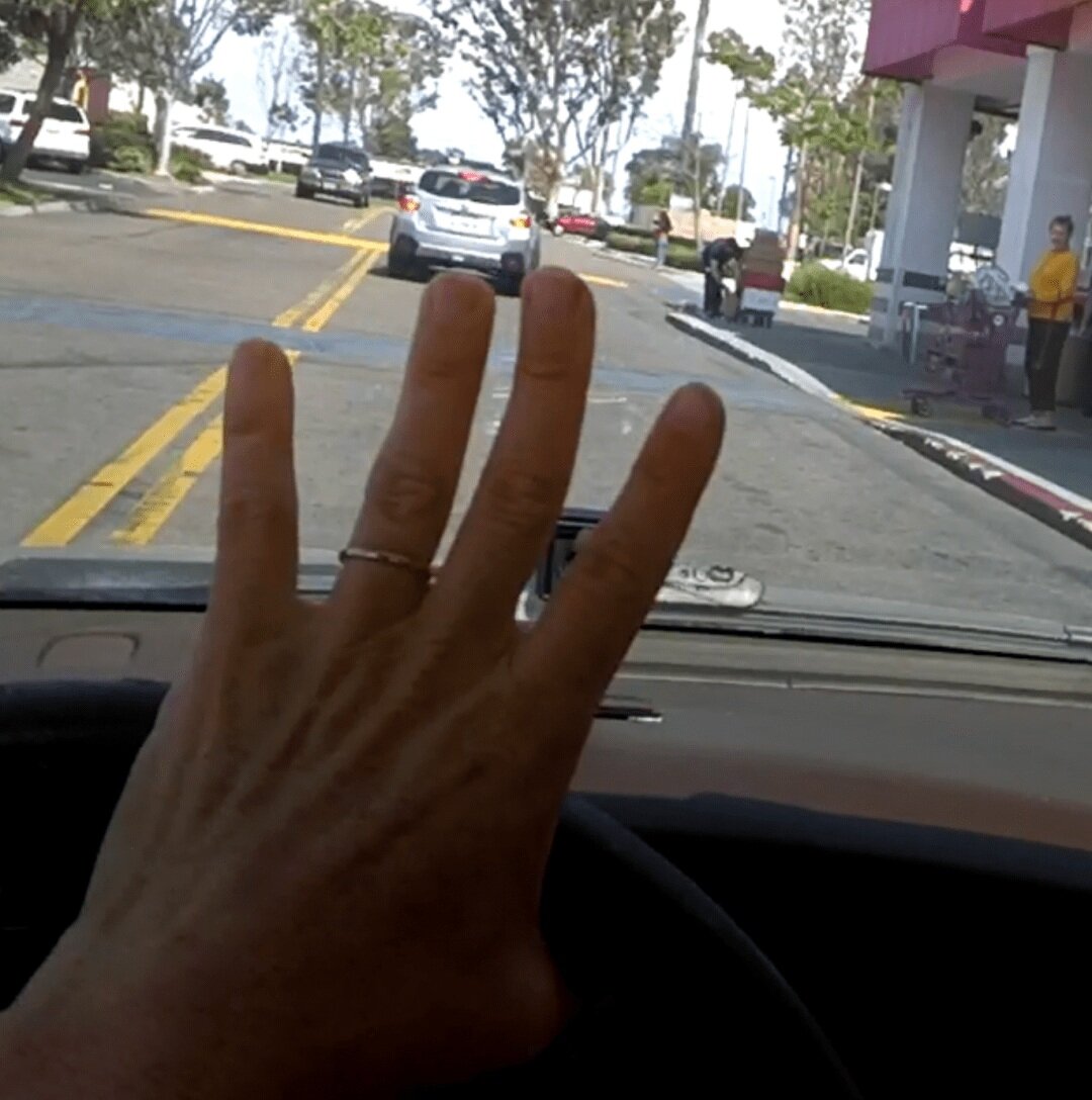 First-person view from inside a car — the driver's hand waves through the windshield to signal a pedestrian that they can cross.