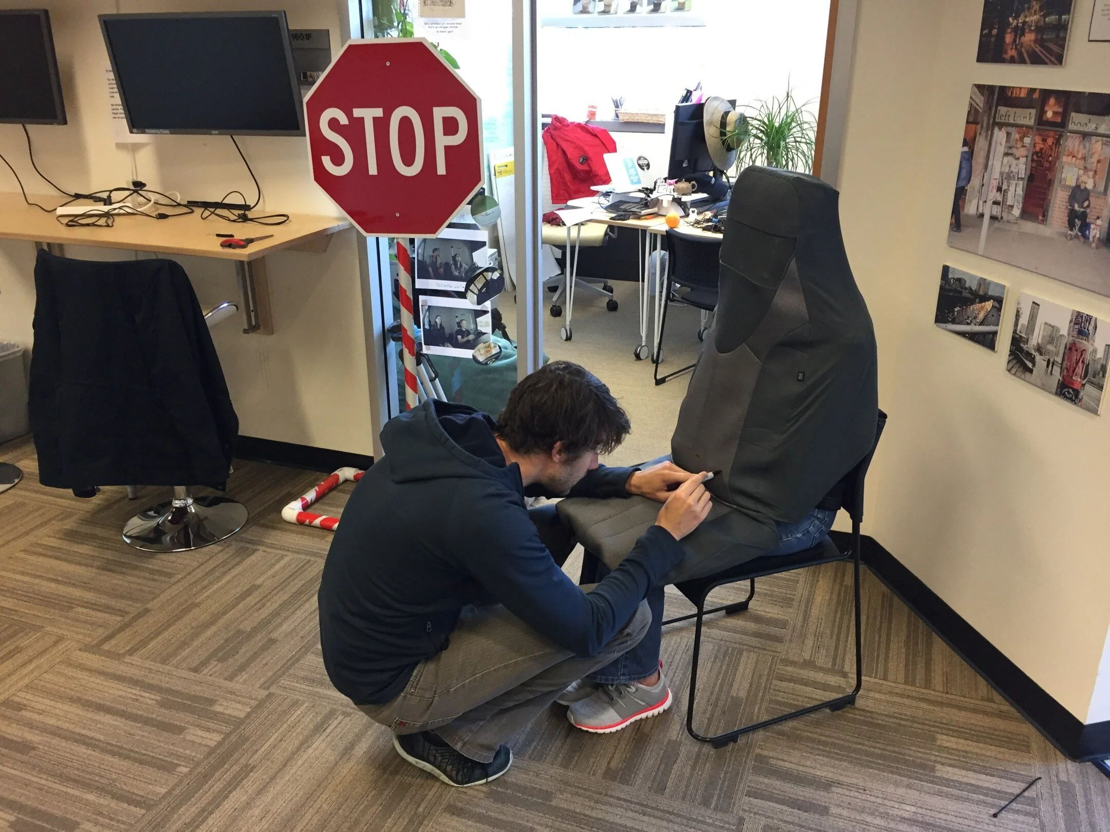 In the lab, a researcher crouches to stitch a grey car-seat cover over a chair, building the seat suit; a STOP sign prop stands behind him.