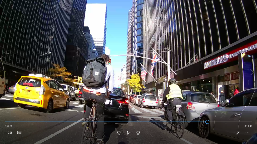 A frame from the handlebar camera on a Manhattan avenue: the expert bicyclist rides in the bike lane alongside a line of cars and a bus, pedestrians crossing ahead.