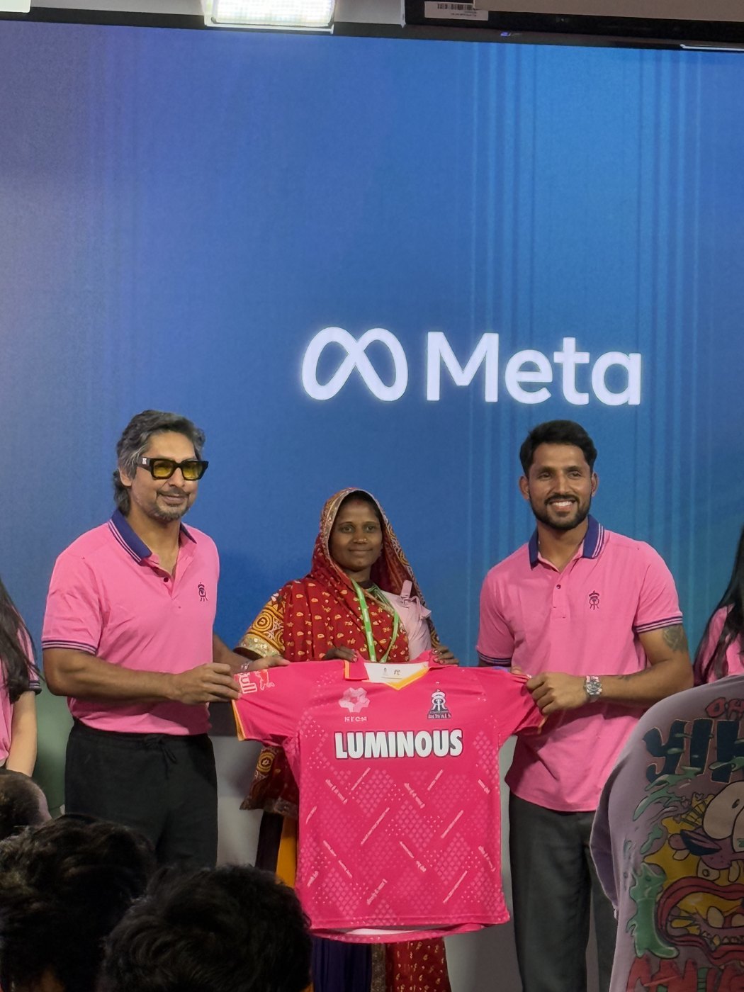 On a stage in front of a large Meta logo, Kumar Sangakkara (left) and Dhruv Jurel (right) of the Rajasthan Royals, in pink team polos, hold up a pink Royals jersey with LUMINOUS across the chest together with a woman in a red-and-gold sari.