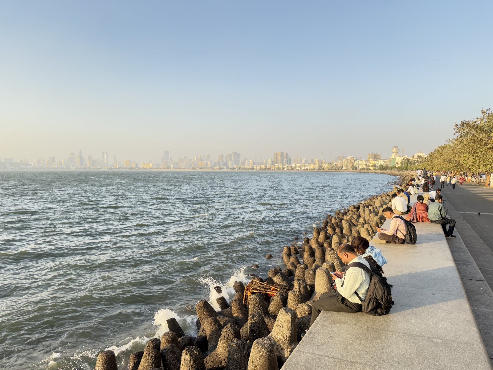 Marine Drive in Mumbai at golden hour: people sit along the sea wall above the tetrapods, the bay and the skyline beyond.