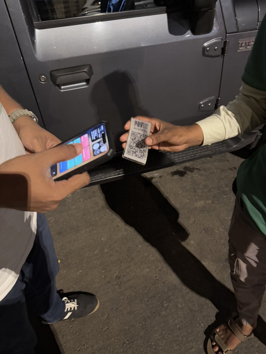 A parking attendant holds out a Paytm “Accepted Here” QR card beside a parked SUV while a customer opens a payment app on their phone.